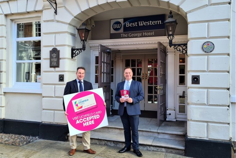 Two people outside The George Hotel holding a Visit Lichfield Card sign and a card.