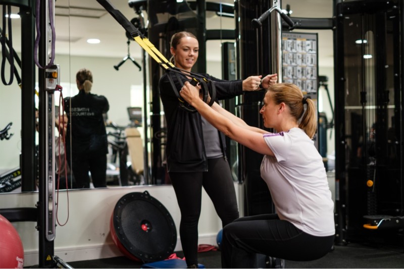 A gym member exercising with suspension straps alongside an instructor